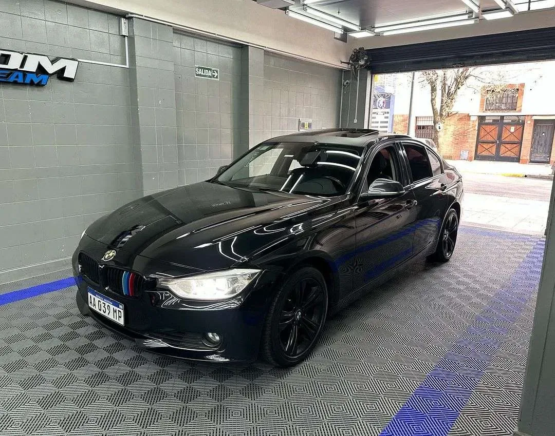 BMW Serie 3 negro dentro del taller ADM TEAM, con el logo del taller en la pared y el piso en damero gris y azul