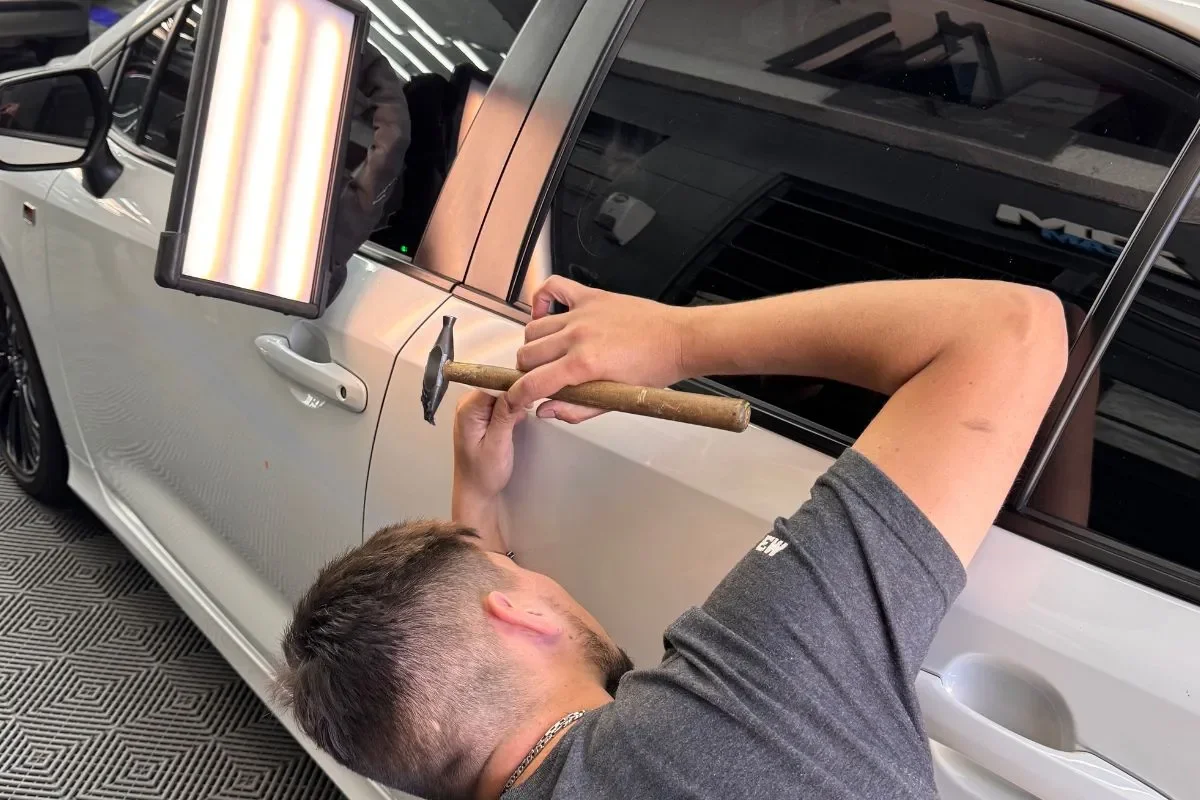 Técnico de ADM TEAM trabajando una abolladura en la puerta de un auto blanco con herramientas de PDR, guiado por la lámpara de reflejo